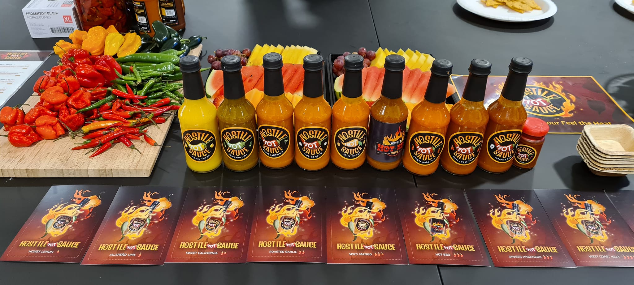 Hot sauce bottles standing side by side with fruit laid out in the background